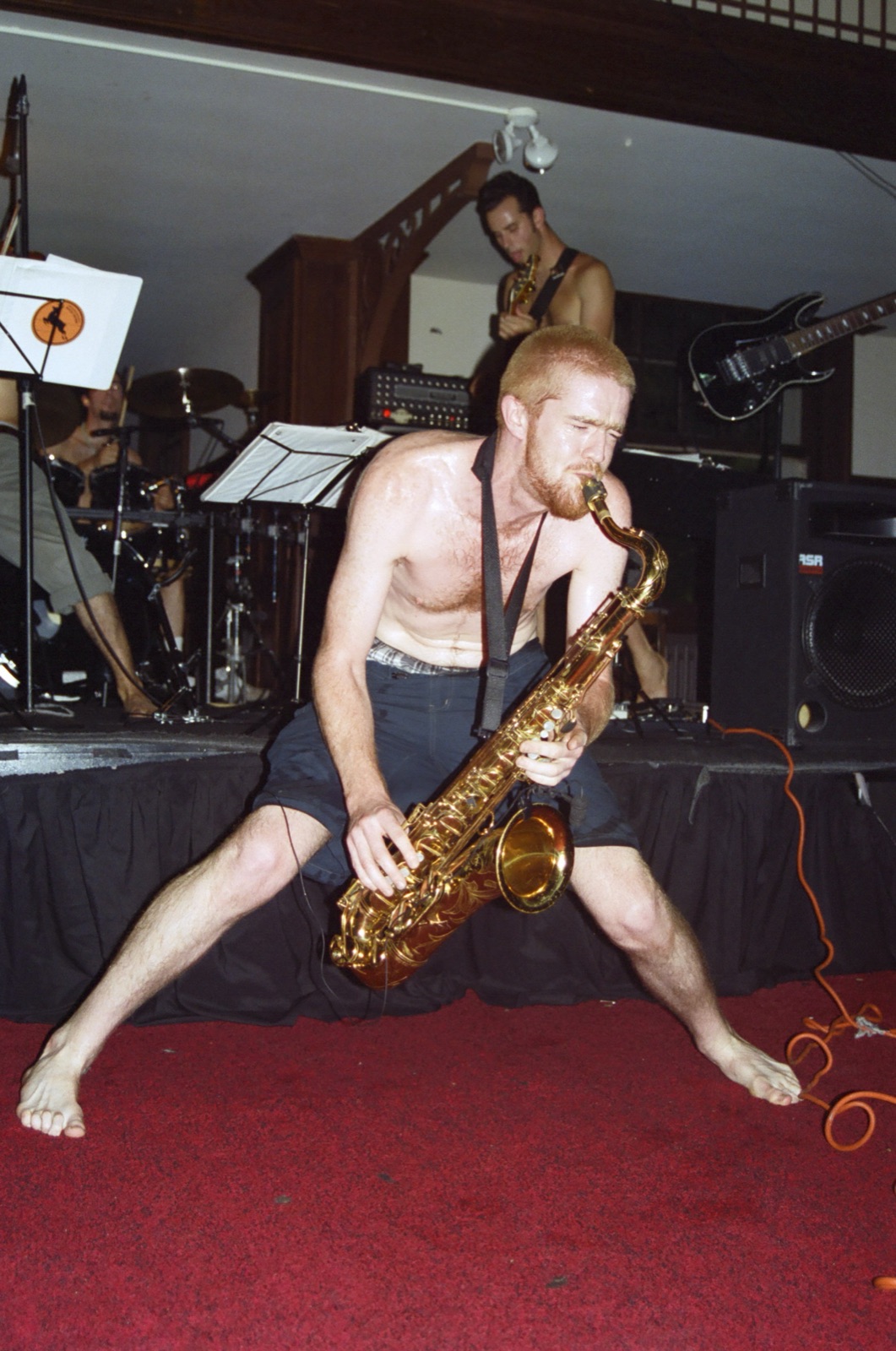 A barefoot shirtless saxophonist crouches in a wide stance on red carpet in front of the stage.